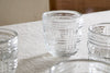The nkuku Kaanch Tumbler - Clear (Set of 4) features embossed check pattern glassware with a thick, sturdy design, displayed on a woven tablecloth with natural light highlighting their craftsmanship and subtle wooden accents in the background.