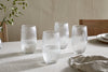 A set of four nkuku Otuba Highball Clear glasses is arranged on a light beige tablecloth, each half-filled with water. Wooden chairs, stacked plates, a cloth napkin, and a small vase of greenery complete the elegant table setting.