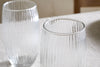 A set of four clear Otuba Highball glasses by nkuku, featuring vertical grooves and gently curved sides, sit on a light fabric surface with natural light emphasizing their elegant ribbed texture.