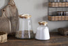 Two nkuku Chaaval Storage Jars with wooden lids sit on a rustic countertop. The larger jar holds granola, the smaller sugar. Wooden boards and a wicker basket complete the organised look, with more jars displayed on a shelf behind.