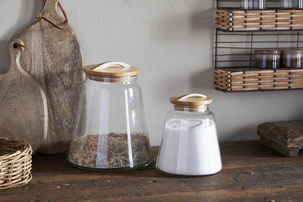 Two nkuku Chaaval Storage Jars with wooden lids sit on a rustic countertop. The larger jar holds granola, the smaller sugar. Wooden boards and a wicker basket complete the organised look, with more jars displayed on a shelf behind.