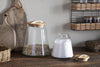 Two nkuku Chaaval Storage Jars with wooden lids sit on a rustic countertop, ideal for kitchen organisation—one filled with brown grains, the other with white powder. Cutting boards, baskets, and more jars are visible in the background.