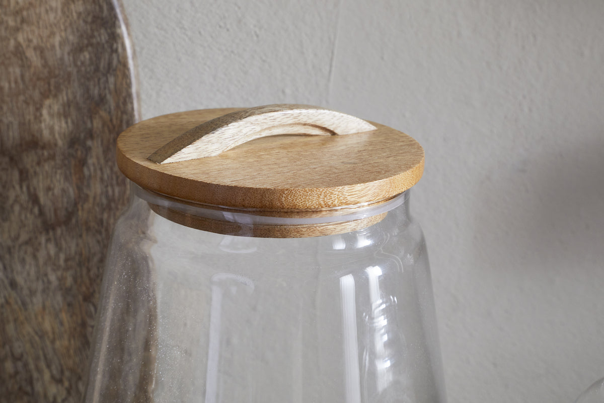 The nkuku Chaaval Storage Jar - Clear features a round, light wood lid with an arched handle. Ideal for kitchen organization, it adds a stylish touch against neutral tones and textured wood backgrounds.