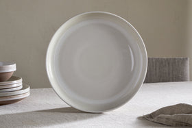 The Edo Dinner Plate - Terracotta by nkuku stands upright on a neutral tablecloth. Stacked everyday dinnerware sits to the left, with a striped chair and light wall creating a calm, minimalist dining scene.