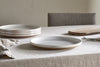 A neutral-toned dining table set with a light linen tablecloth, stacked ceramic plates, an nkuku Edo Dinner Plate in terracotta front and center, a wooden cutting board, silverware, and folded napkin highlights minimalist style. An upholstered chair sits behind.