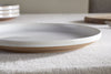The nkuku Edo Dinner Plate - Terracotta features a white glazed ceramic surface with a brown unglazed rim, displayed on a textured beige tablecloth. In the background, stacked plates, a wooden board, and a fork appear softly out of focus.