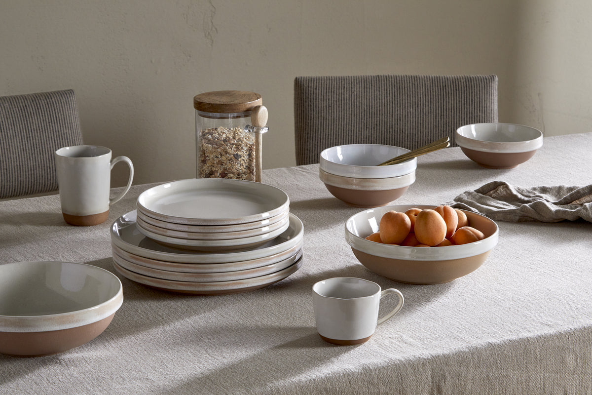 A beige tablecloth covers a dining table set with nkuku's Edo Dinner Plate in Terracotta, plus stacked ceramic bowls and mugs. A bowl of apricots, a jar with wooden lid, and a folded napkin complete the minimalist look.