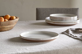 A stack of ceramic plates and the nkuku Edo Side Plate - Terracotta rest on a textured beige tablecloth. To the left, a wooden bowl holds apricots, while a gray napkin completes this elegant minimalist tableware arrangement.