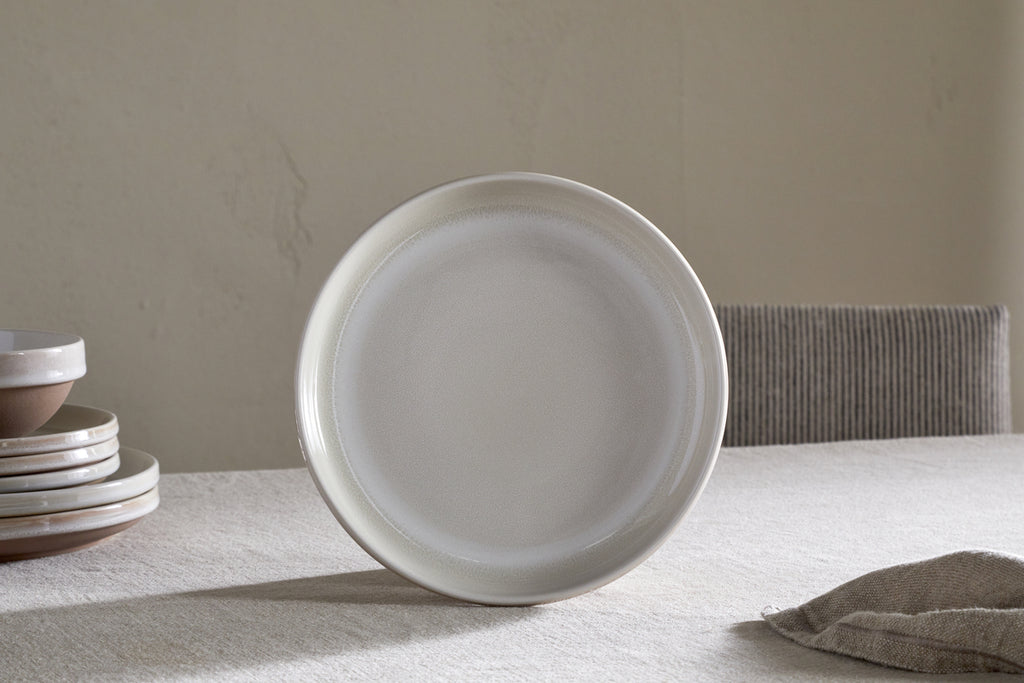 The Edo Side Plate - Terracotta by nkuku stands upright on a beige tablecloth, with stacked plates and bowls to the left and a folded textured napkin on the right, creating a minimalist aesthetic in soft lighting.