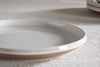 A close-up of the nkuku Edo Side Plate - Terracotta, featuring a beige and white glaze with a subtle brown rim and glossy finish, displayed on textured fabric. Another similar plate is partially visible in the background.