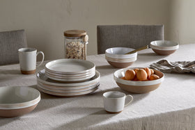 A table set with minimalist beige ceramic plates, bowls, and cups—some stacked. A large nkuku Edo Cereal Bowl in Terracotta holds apricots. There’s a glass jar of oats, striped napkin, and neutral chairs on a light tablecloth.