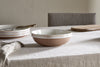 A handcrafted nkuku Edo Pasta Bowl - Terracotta with a white interior and terracotta exterior rests on a light linen tablecloth, surrounded by minimalist tableware—stacked plates, fork, wooden board—and a gray upholstered chair in a simple setting.