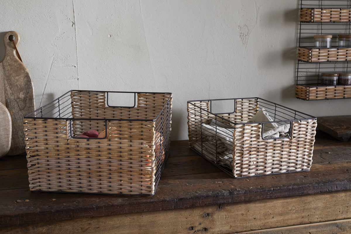 Two Montali Iron & Rattan Storage Baskets by nkuku sit on a rustic wooden table, one holding a pink cloth and the other a white cloth. Behind them, cutting boards and shelves with jars complete the scene.