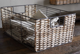The Montali Iron & Rattan Storage Basket by nkuku, with a metal frame and natural rattan sides, sits on a wooden surface holding neatly folded beige towels. Open handles add to its rustic look; another basket hangs on the wall.