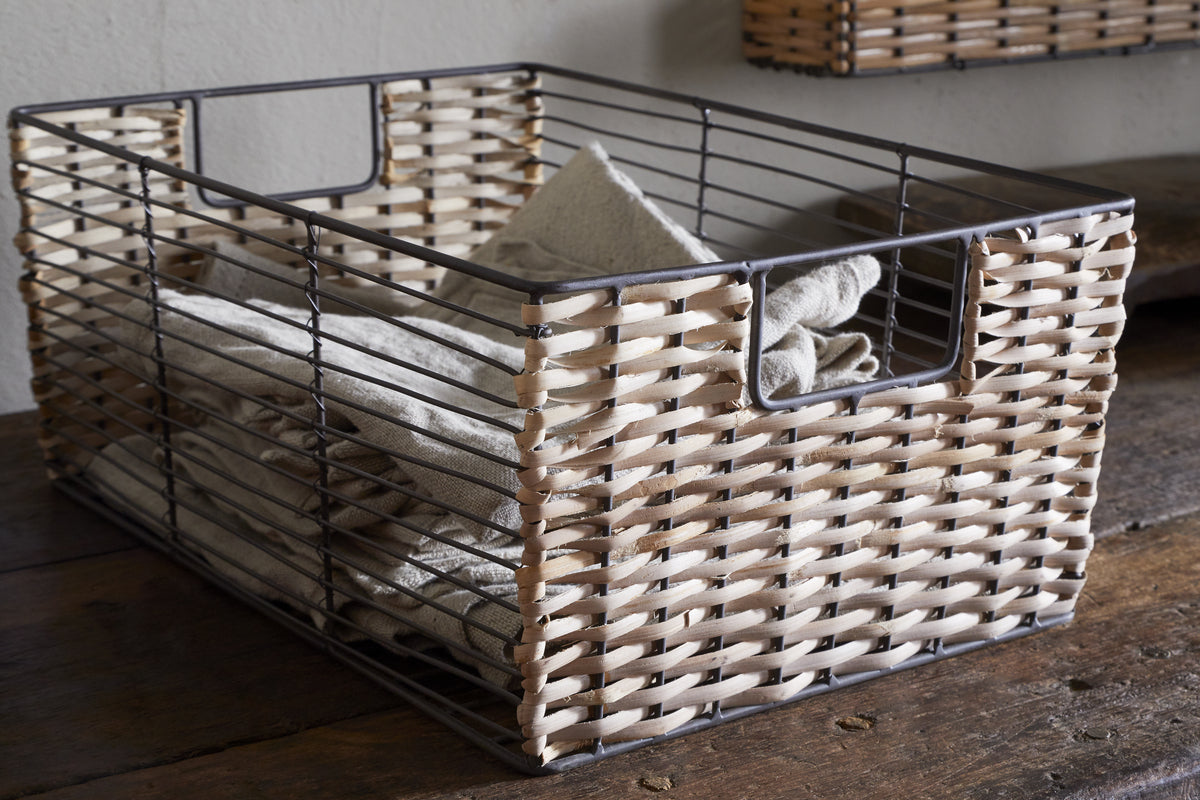 The Montali Iron & Rattan Storage Basket by nkuku, with a metal frame and natural rattan sides, sits on a wooden surface holding neatly folded beige towels. Open handles add to its rustic look; another basket hangs on the wall.