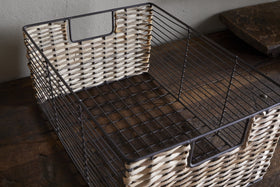 The Montali Iron & Rattan Storage Basket by nkuku, in natural white and brown with a metal frame, open handles, and grid-patterned base, rests on a dark wooden surface against a light wall.