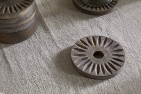 The Eisha Mango Wood Coasters by nkuku, in dark brown with hand-carved sunburst design, rest on textured beige fabric. A stacked set of six and a partially visible coaster share the elegant detailing.
