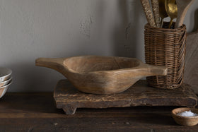 The Naashta Handled Serving Bowl – Natural by nkuku, with its rustic wood and twin handles, sits on a textured board. A woven utensil holder filled with kitchen tools and salt stands right; stacked white bowls rest left, creating a cozy vintage kitchen vibe.