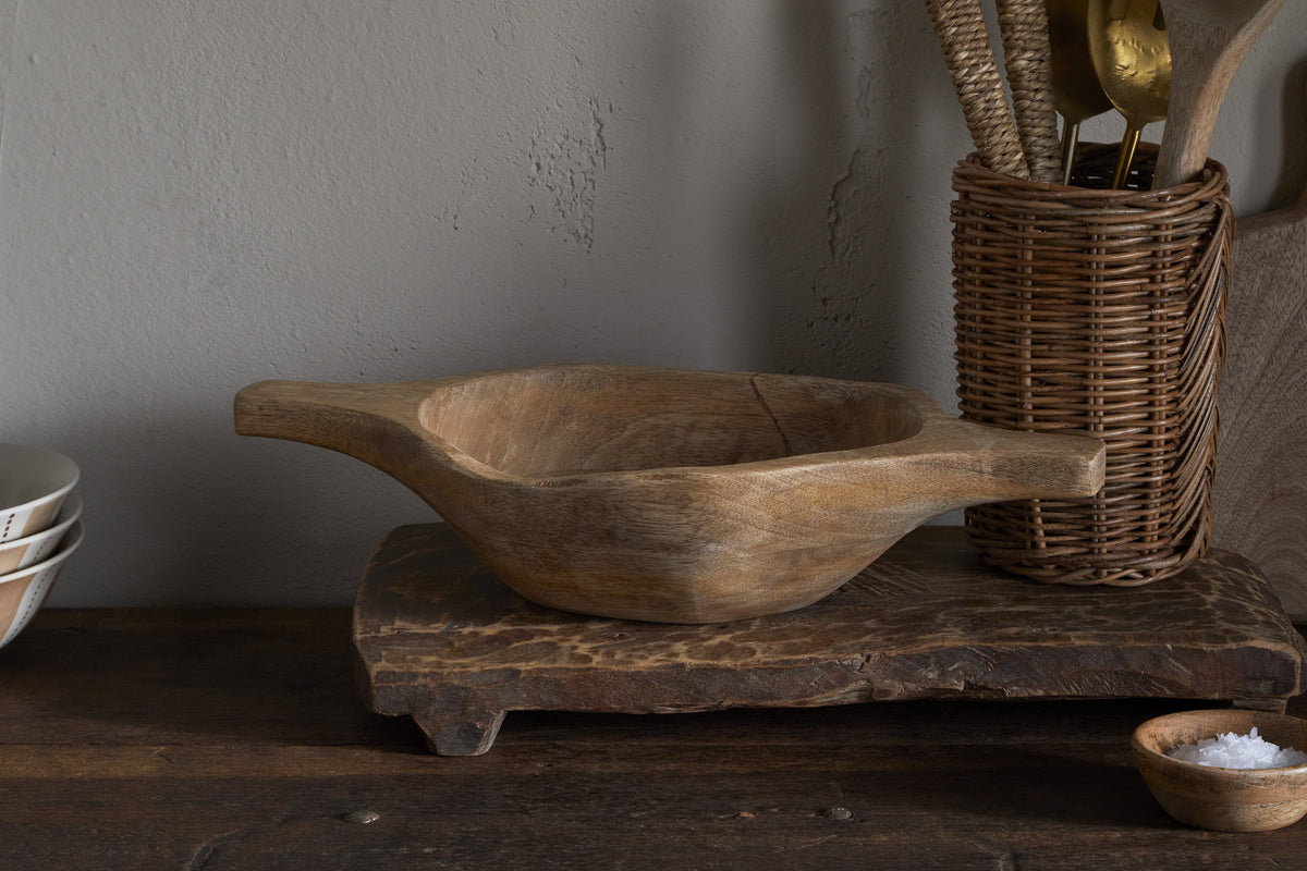 The Naashta Handled Serving Bowl – Natural by nkuku, with its rustic wood and twin handles, sits on a textured board. A woven utensil holder filled with kitchen tools and salt stands right; stacked white bowls rest left, creating a cozy vintage kitchen vibe.