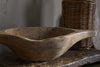 The Naashta Handled Serving Bowl - Natural by nkuku, a rustic hand-carved piece, rests on wood beside a woven wicker container, creating a cozy kitchen vibe with earthy tones and textured walls in the background.