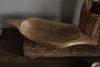 The Naashta Handled Serving Bowl - Natural by nkuku, an oval-shaped, hand-carved wooden bowl with handles, rests empty on a rustic kitchen surface. A woven utensil basket stands nearby against a neutral textured wall.