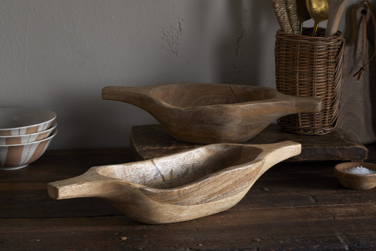 Two Naashta Handled Serving Bowls by nkuku sit on a dark dining table—one in the foreground, one behind it on a wooden board. Around them are stacked white bowls, a wicker utensil holder with golden utensils, and a small salt dish.