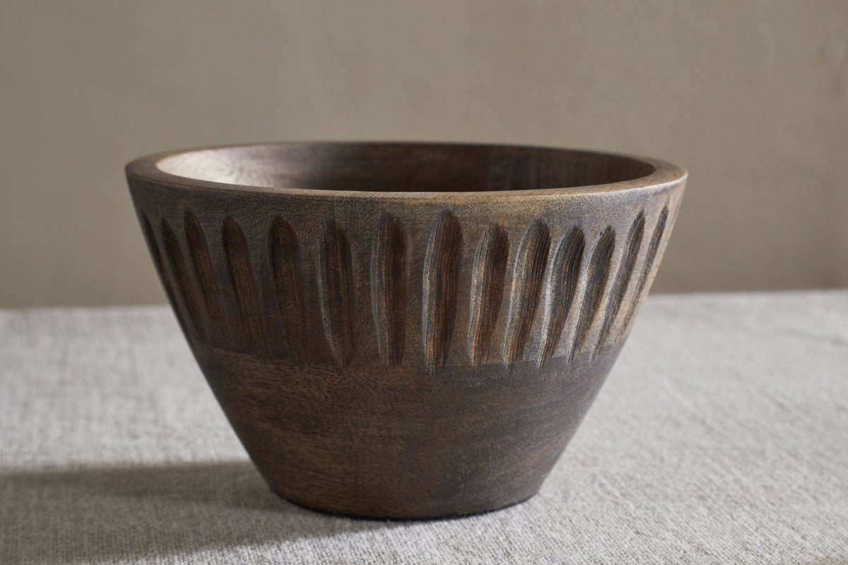 The nkuku Eisha Serving Bowl - Brown, featuring a wide rim and hand-carved leaf-like pattern, rests on textured fabric against a plain beige background. Its dark wood matte finish adds elegance to any setting.