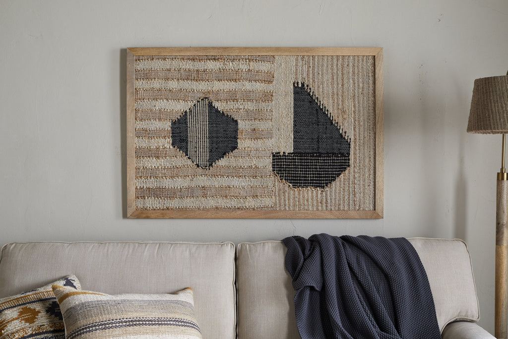The Kiran Large Textured Wall Art - Natural & Dark Blue by nkuku hangs above a light gray sofa with textured pillows and a dark blue knitted throw, adding cozy modern style. A standing lamp appears to the right.