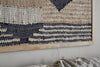 A close-up of nkuku's Birala Large Textured Wall Art in natural and dark blue hangs on a light wall. Black, cream, and brown abstract geometric patterns stand out. Below, a beige textile is draped over the couch.