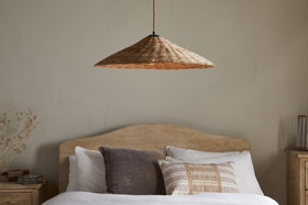 A cozy bedroom features a bed with a wooden headboard, white sheets, and cushions. Above it hangs nkuku’s Sangu Rattan Pendant in natural, casting soft light. Light beige walls, nightstands, and dried flowers add warmth to the space.