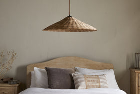 A minimalist bedroom features a light wood bed with a curved headboard, white bedding, and textured pillows. Above the bed hangs nkuku’s Sangu Rattan Pendant in natural, casting soft light. Light beige walls and dried flowers decorate the wooden nightstand.