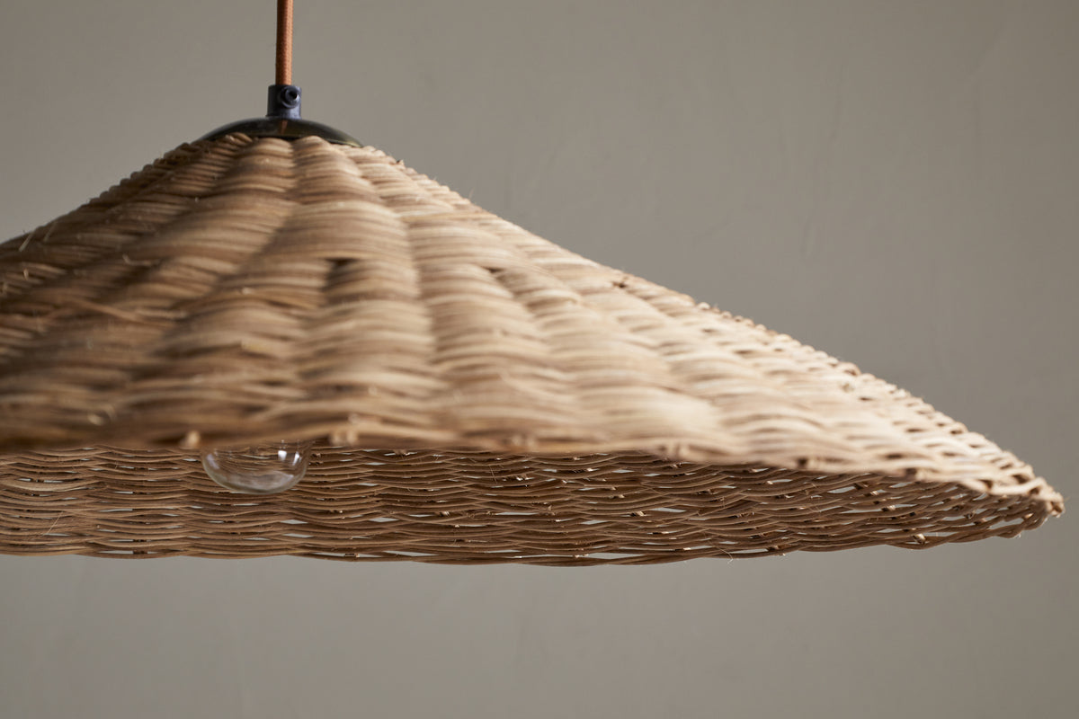 A close-up of the Sangu Rattan Pendant - Natural by nkuku shows its wide, conical woven design. An exposed bulb below casts soft light, highlighting the natural wicker texture and fine craftsmanship against a plain background.