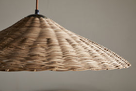 A close-up of the nkuku Sangu Rattan Pendant in Natural shows its large, conical wicker shade hanging from the ceiling. Soft light passes through the woven texture, casting gentle shadows on a neutral wall.