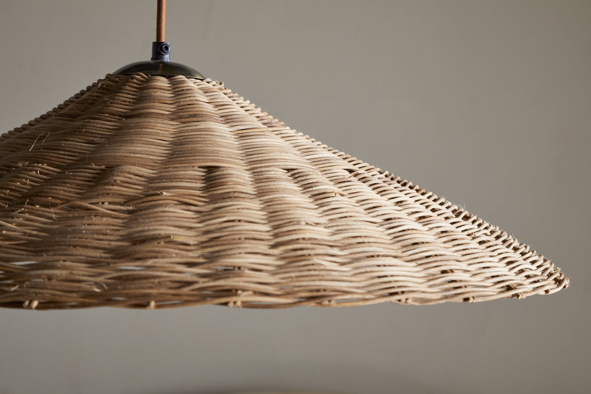 A close-up of the nkuku Sangu Rattan Pendant in Natural shows its large, conical wicker shade hanging from the ceiling. Soft light passes through the woven texture, casting gentle shadows on a neutral wall.