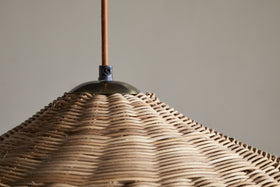Close-up of the Sangu Rattan Pendant - Natural by nkuku, featuring a conical, handwoven rattan shade suspended from a brown fabric cord. The intricate pattern and soft light showcase the craftsmanship and natural beauty of this wicker pendant.
