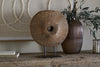 The Cala Reclaimed Wood Objet on Stand - Natural by nkuku, a round rustic sculpture with a center hole on a black metal stand, rests on a table beside a ribbed brown vase with branches, a candle, books, and circular artwork in the background.