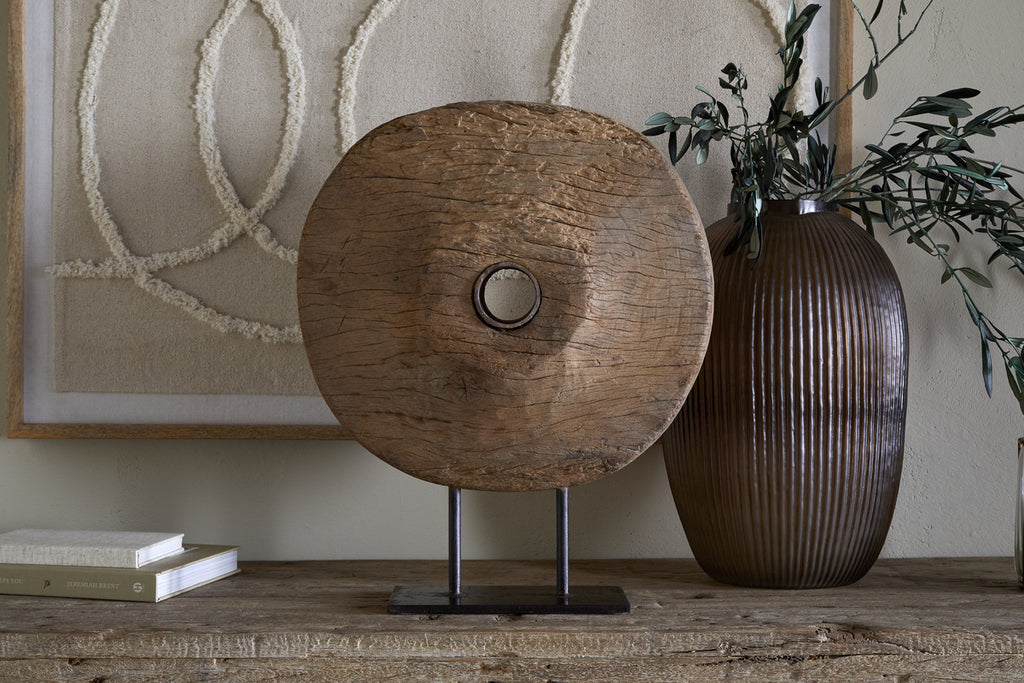 The Cala Reclaimed Wood Objet on Stand - Natural by nkuku features a rustic wooden disc with a central hole on a black metal base, displayed beside a ribbed brown vase, green branches, abstract wall art, and two stacked books.