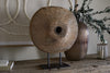 The nkuku Cala Reclaimed Wood Objet on Stand – Natural, a rustic round sculpture with a central hole, is displayed on a weathered wood surface beside a ribbed bronze-toned vase with leafy branches for an elegant décor arrangement.