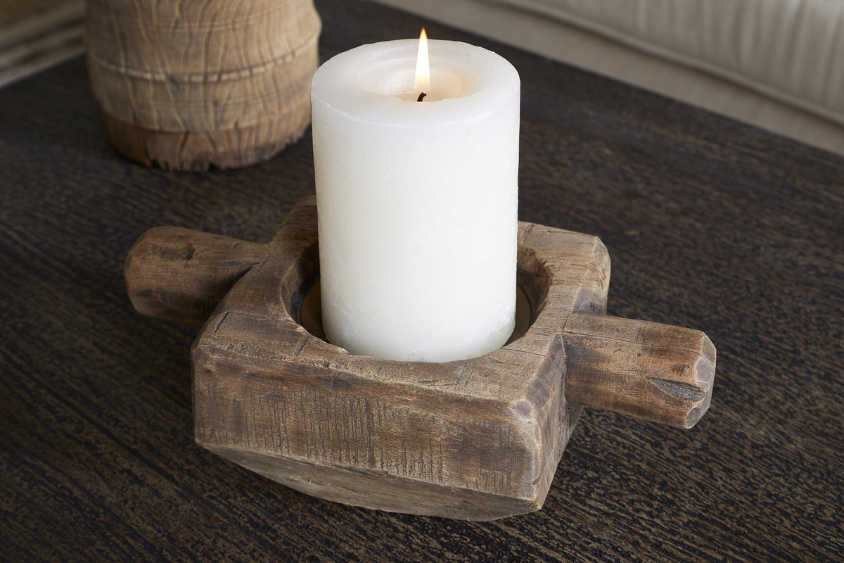 A lit white pillar candle sits in the nkuku Apu Reclaimed Wood Traditional Candle Holder - Natural, featuring two short wooden rod handles, set on a dark textured wooden surface. A blurred object appears in the background.