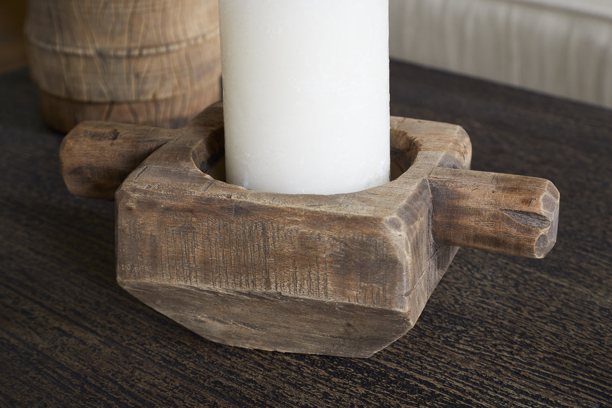 The Apu Reclaimed Wood Traditional Candle Holder - Natural by nkuku features a thick white candle, highlighting the natural wood grain and texture, and rests on a dark wooden surface with another wooden object blurred in the background.