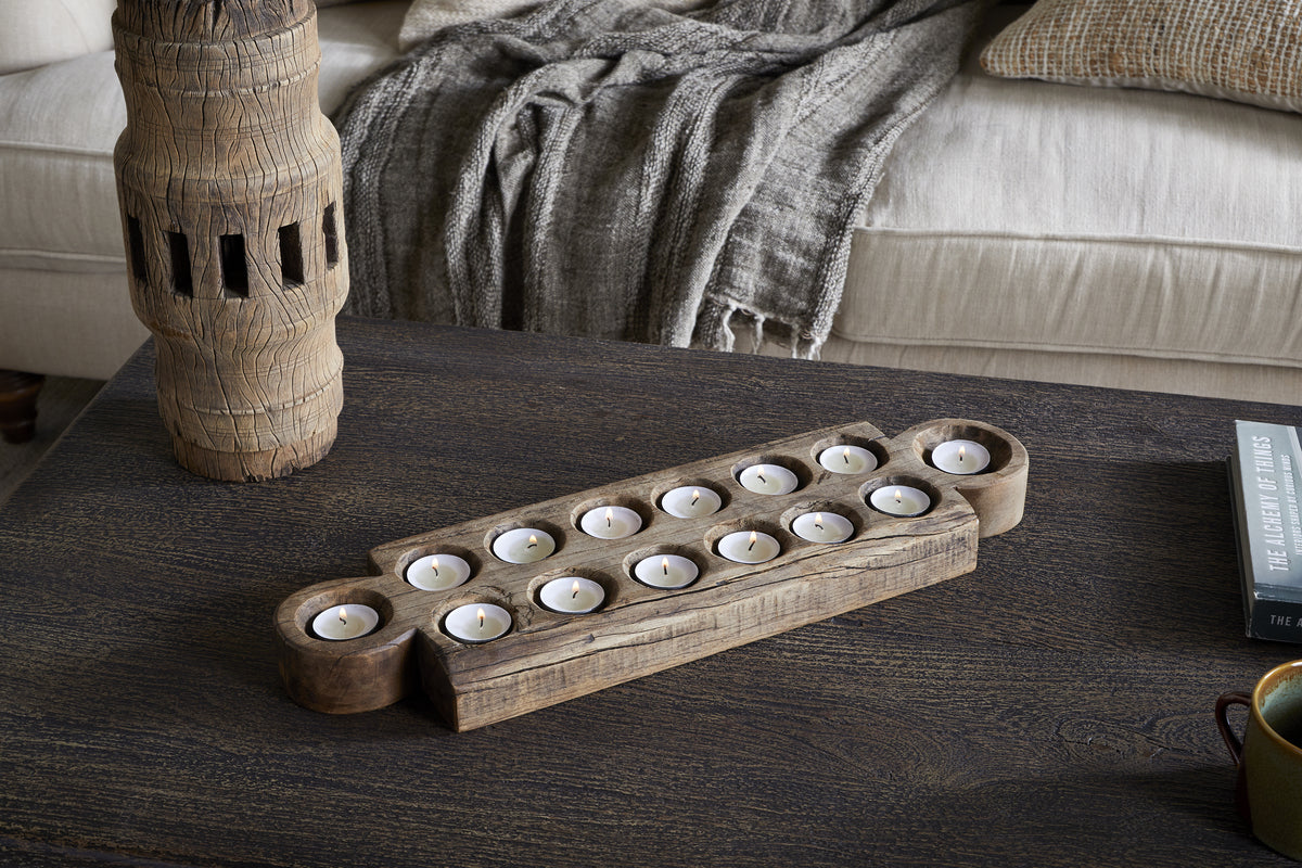 The nkuku Makala Reclaimed Wood Tealight Holder - Natural, with fourteen glowing candles, rests on a dark wooden coffee table. A tall carved decor piece, grey textured throw on a beige sofa, and The Legend of Lythmus book complete the look.