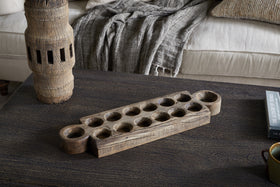 The Makala Reclaimed Wood Tealight Holder - Natural by nkuku, with carved circular pits, sits on a dark coffee table near a weathered accent, gray throw on a beige couch, and a black book titled THE LEGACY OF TITHES—a striking decor piece.
