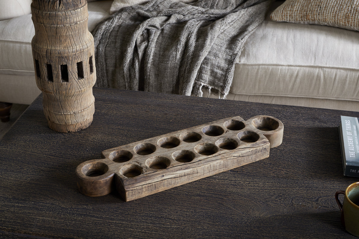 The Makala Reclaimed Wood Tealight Holder - Natural by nkuku, with carved circular pits, sits on a dark coffee table near a weathered accent, gray throw on a beige couch, and a black book titled THE LEGACY OF TITHES—a striking decor piece.