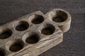A close-up of the nkuku Makala Reclaimed Wood Tealight Holder - Natural, featuring eight round tealight slots and one larger semi-circular end slot. This decorative piece has a textured, weathered finish and rests unused on a dark, grainy surface.