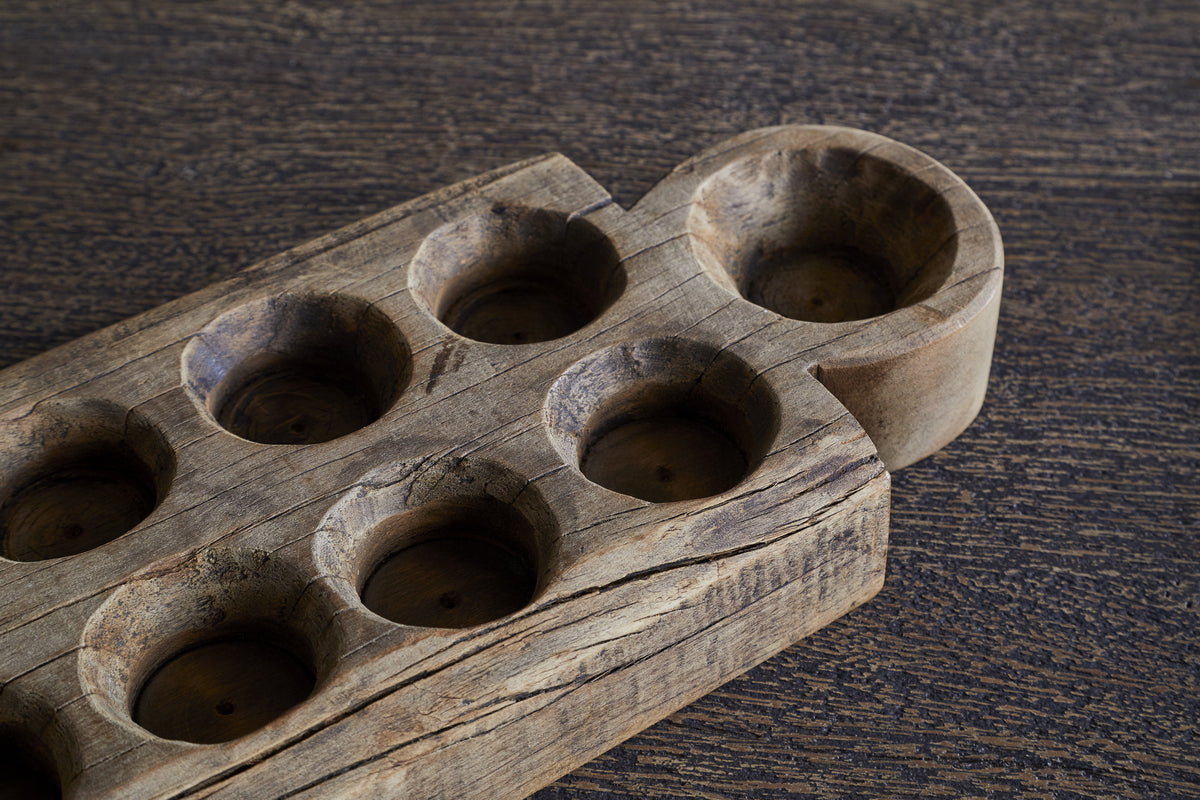 A close-up of the nkuku Makala Reclaimed Wood Tealight Holder - Natural, featuring eight round tealight slots and one larger semi-circular end slot. This decorative piece has a textured, weathered finish and rests unused on a dark, grainy surface.