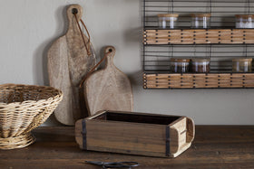 A rustic kitchen features two cutting boards, a wicker basket, and the nkuku Dudha Reclaimed Brick Box - Natural used as a decorative storage container. A wire shelf with spice jars and nearby scissors complete the scene.