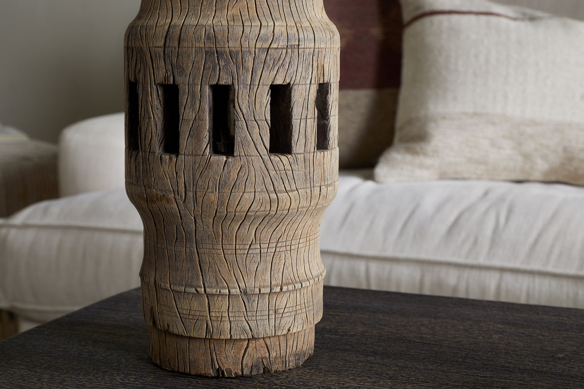 The Mvua Reclaimed Wood Pillar Candle Holder - Natural by nkuku, featuring vertical rectangular cutouts, rests on a dark wooden table with a beige couch and textured cream and taupe cushions in the background for a cozy touch.