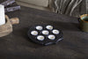 The nkuku Maji Stone Multi Tealight Holder in Antique Black with seven white candles sits on a dark wooden table beside a green ceramic mug, a large white pillar candle on a wooden tray, and a gray textured blanket over a beige couch.