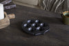 A dark wooden table displays the nkuku Maji Stone Multi Tealight Holder in Antique Black with seven tealights. Nearby are a green ceramic mug, a wooden tray holding a white candle, and a gray textured blanket on a beige sofa.