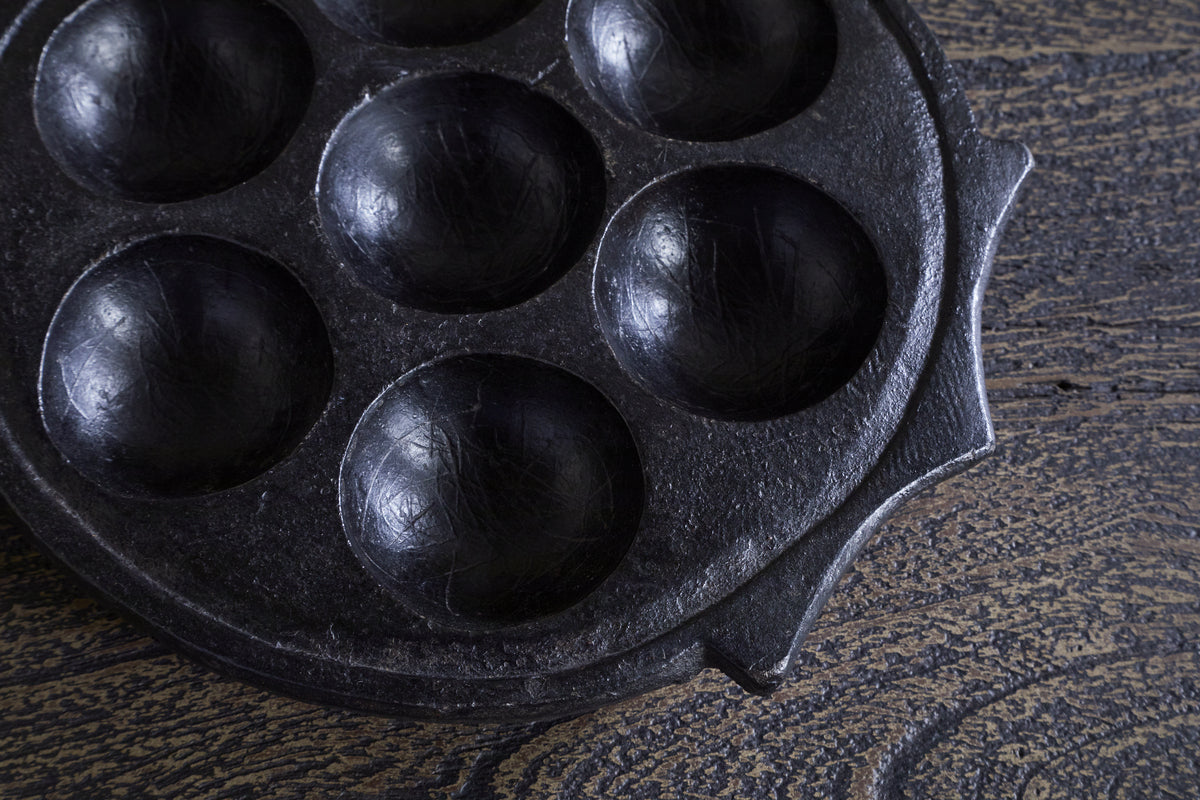 A close-up of the nkuku Maji Stone Multi Tealight Holder in Antique Black, featuring seven round wells, sits on textured wood. This functional décor piece has a seasoned, slightly worn surface for a distinctive look.